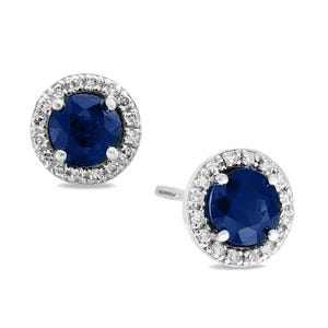 A pair of stud earrings featuring round blue gemstones at the center, surrounded by a halo of small clear stones set in a silver-toned metal.