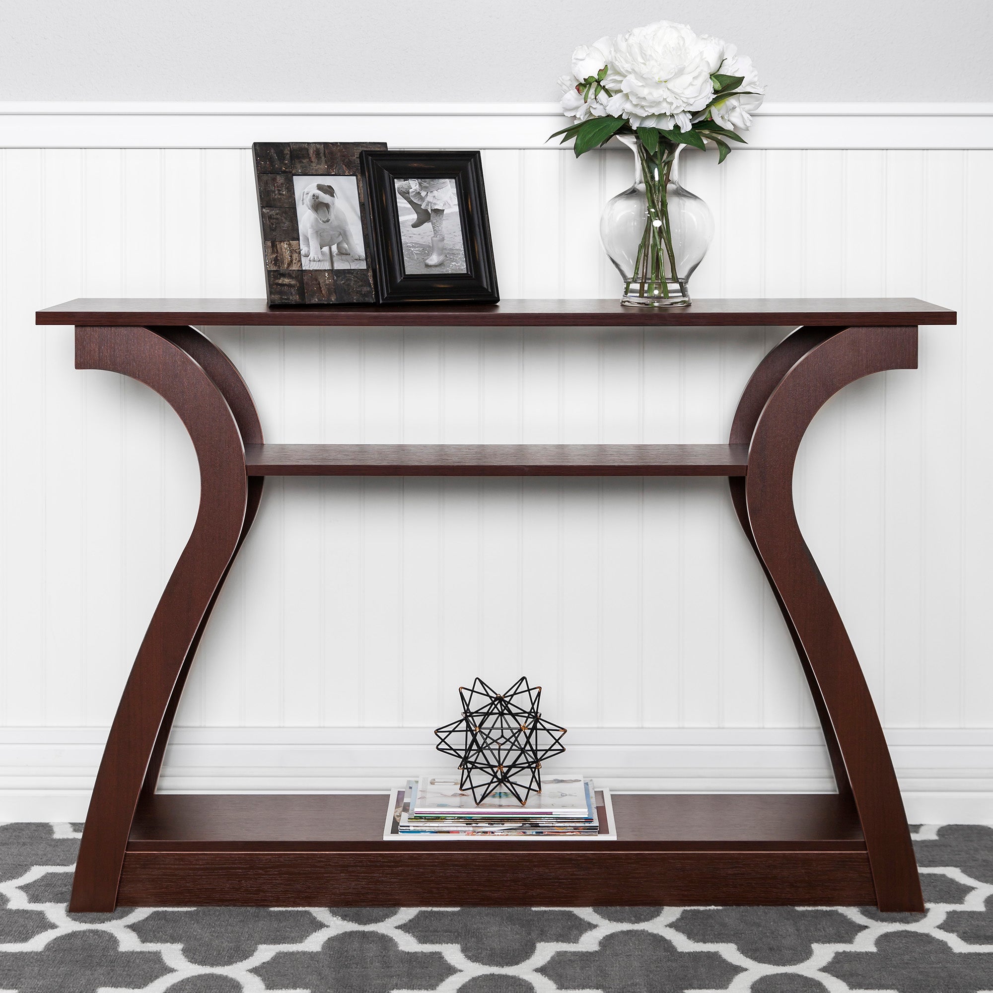 A 47-inch wide wooden accent table with curved legs features a lower shelf, displaying a geometric ornament and magazines, and a top displaying a framed photograph and a vase of white flowers.