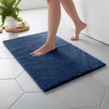 A person steps onto a plush navy blue bath mat placed on hexagonal white tile flooring. A white planter with green ferns is visible in the background.