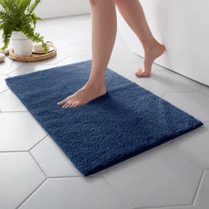 A person steps onto a plush navy blue bath mat placed on hexagonal white tile flooring. A white planter with green ferns is visible in the background.
