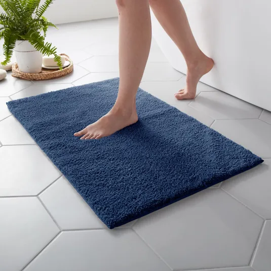 A person steps onto a plush navy blue bath mat placed on hexagonal white tile flooring. A white planter with green ferns is visible in the background.