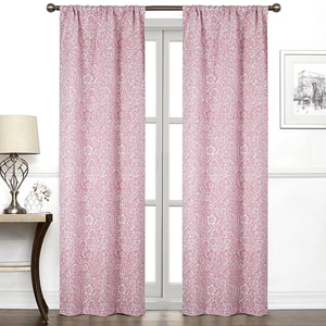 Pink curtains with a white paisley pattern hang on a rod in front of a window. The room features a table with a lamp and a small potted plant, along with framed wall art.