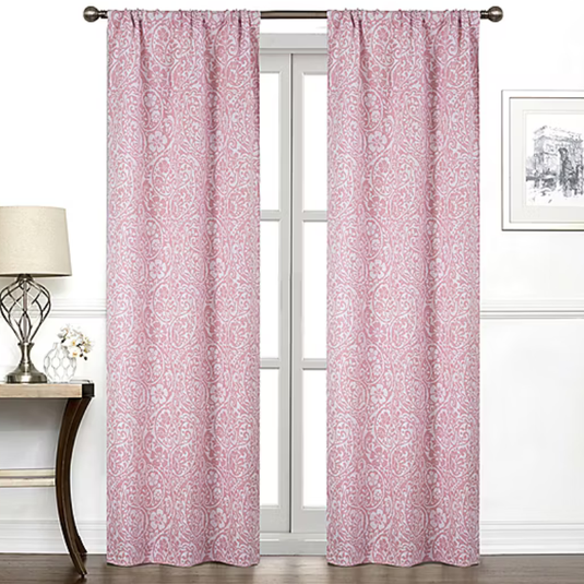 Pink curtains with a white paisley pattern hang on a rod in front of a window. The room features a table with a lamp and a small potted plant, along with framed wall art.
