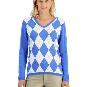 A woman models a blue and white argyle V-neck sweater with long sleeves, featuring a large diamond pattern on the front. She pairs it with light-colored pants.