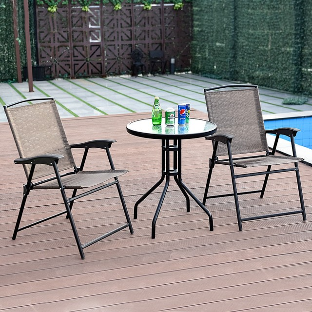 Two outdoor folding chairs are positioned around a small round glass table with metal legs. On the table are a bottle and cans of beverages. The setting is on a wooden deck near a pool.