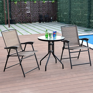 Two outdoor folding chairs are positioned around a small round glass table with metal legs. On the table are a bottle and cans of beverages. The setting is on a wooden deck near a pool.