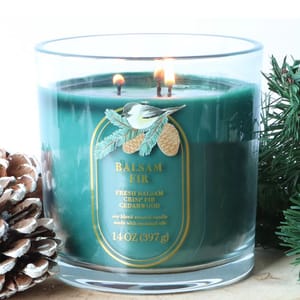 A green 14 oz balsam fir-scented candle featuring fresh balsam, crisp fir, and cedarwood, contained in a clear glass jar, surrounded by pinecones and greenery.