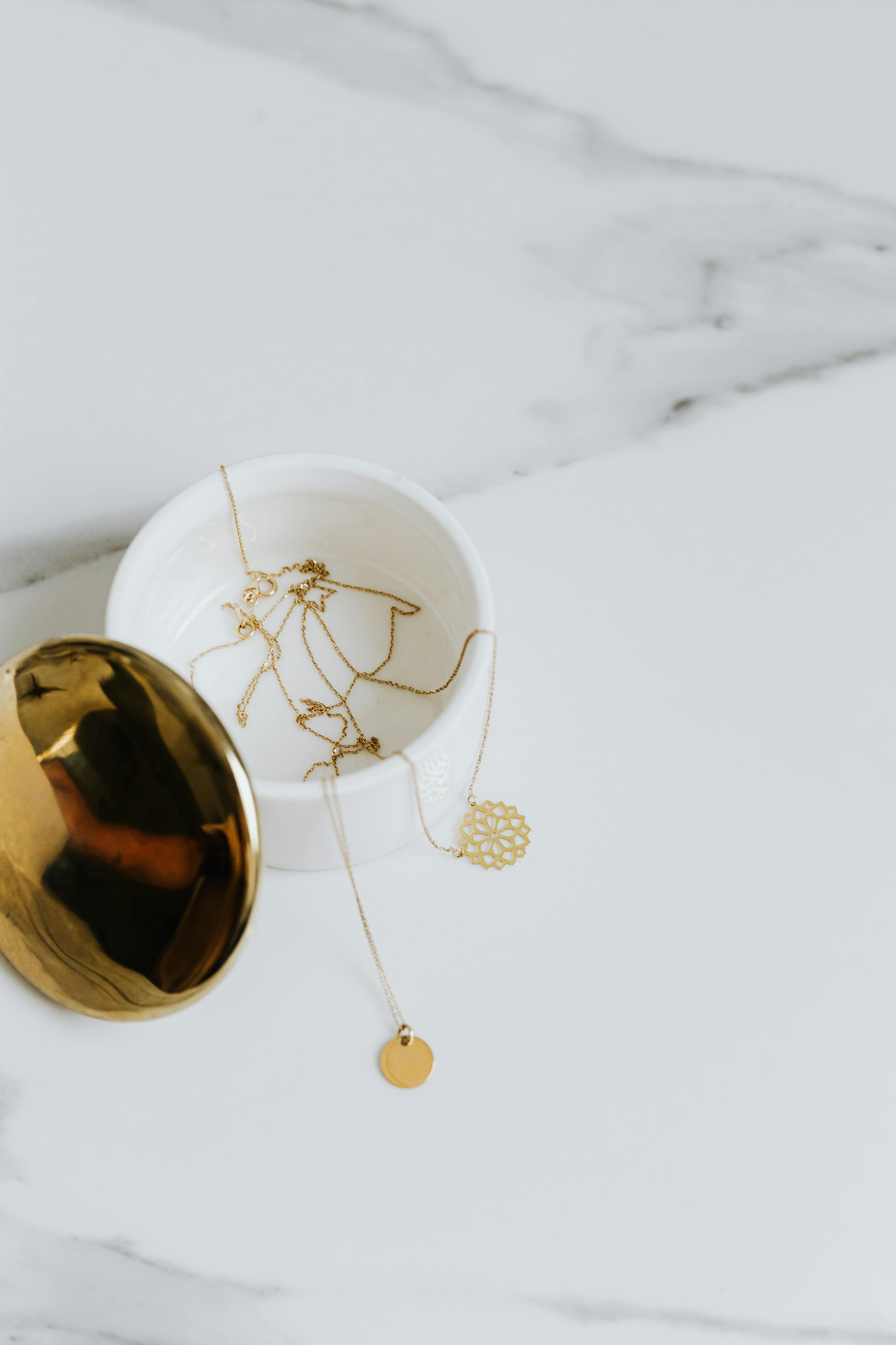 A gold necklace with a circular pendant draping out of an open white circular container on a marble background.