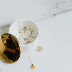 A gold necklace with a circular pendant draping out of an open white circular container on a marble background.