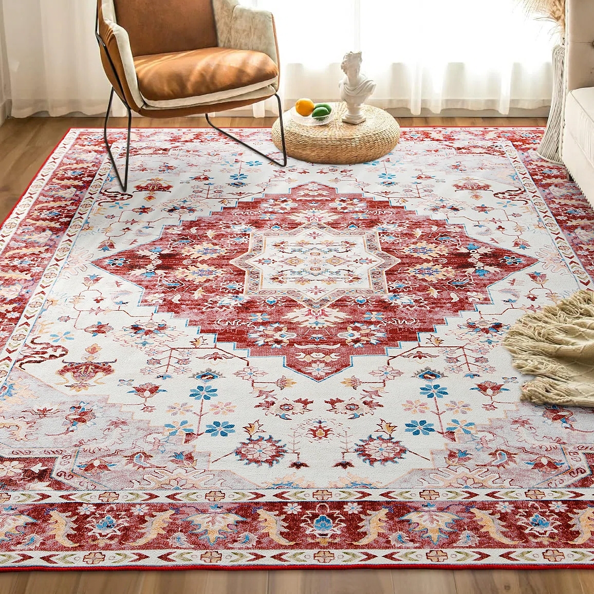 A 5' x 8' machine washable area rug featuring a red, blue, and white intricate pattern with floral and geometric designs, placed in a cozy room setting with a brown armchair and textured pouf.