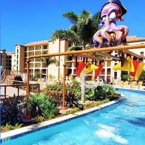 A lazy river pool with tropical foliage features a pirate-themed octopus structure, located in a resort area with multiple buildings in the background.