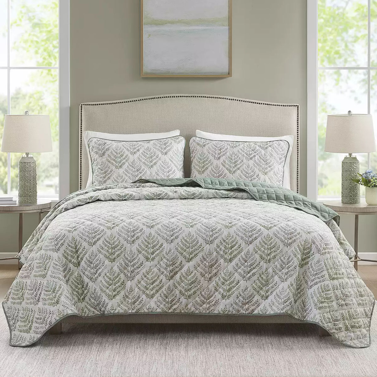 A bed is covered with a quilt and matching pillow shams featuring a green fern pattern. Two bedside tables hold matching lamps with decorative bases, against a backdrop of light-colored walls and windows.