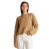 A beige, 100% organic cotton cable knit sweater featuring intricate knit patterns and a relaxed fit, worn by a model with long sleeves and a round neckline, paired with white trousers.