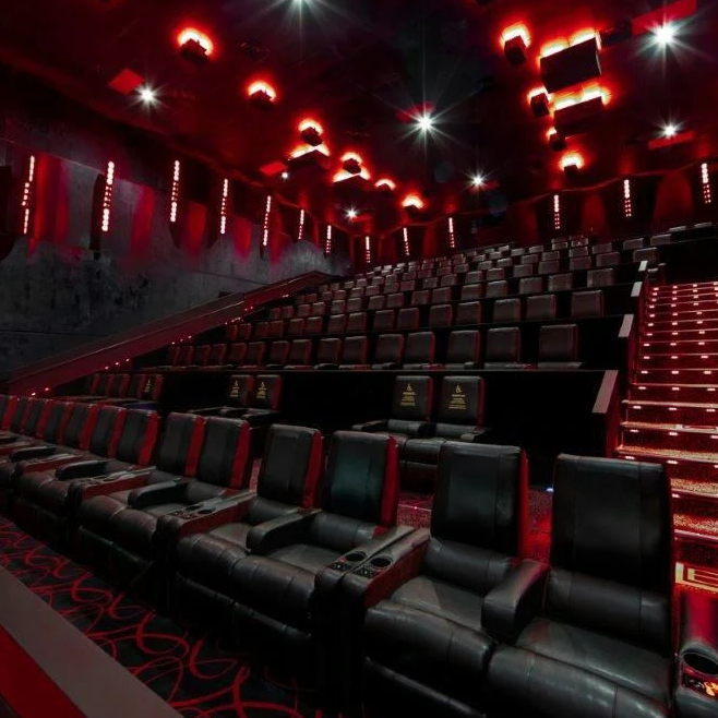 A modern movie theater with black leather recliner seats and red ambient lighting. The auditorium features tiered seating and illuminated aisle steps.
