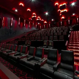 A modern movie theater with black leather recliner seats and red ambient lighting. The auditorium features tiered seating and illuminated aisle steps.