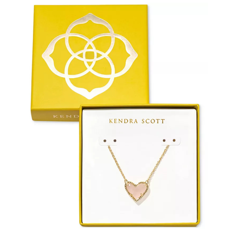 Kendra Scott Boxed Heart Necklace features a pink heart-shaped pendant on a gold chain, packaged in a yellow box with a floral design.