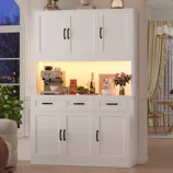 White kitchen cabinet with black handles featuring an espresso machine, bottles, a plant, and a cake with flowers on top. There's a view of a window with a plant in the corner and a cozy chair with a blanket.