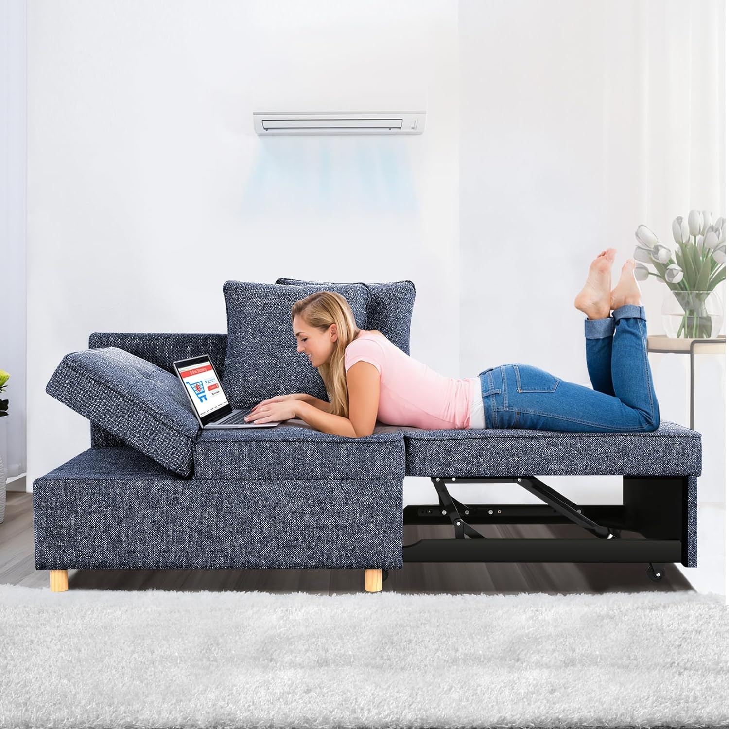 A woman is lying on a denim-textured sofa with an extendable seating area, using a laptop placed on the cushion.