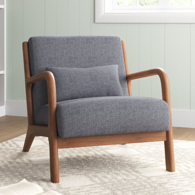 A mid-century solid wood accent chair with a curved wooden frame and grey upholstered seat and back, featuring a rectangular lumbar pillow. It's set on a patterned rug in a light-colored room, offering a retro yet modern appearance.