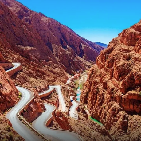 A winding road weaves through a dramatic rocky canyon with reddish cliffs on both sides, under a clear blue sky.
