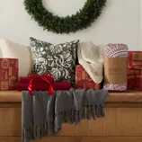 Holiday display with a green wreath, red ribbon, red-patterned gift boxes, a green and white ornamental pillow, cream knit pillow and blanket, a gray fringe throw, red ribboned sheet set, and a reversible patterned quilt set roll.