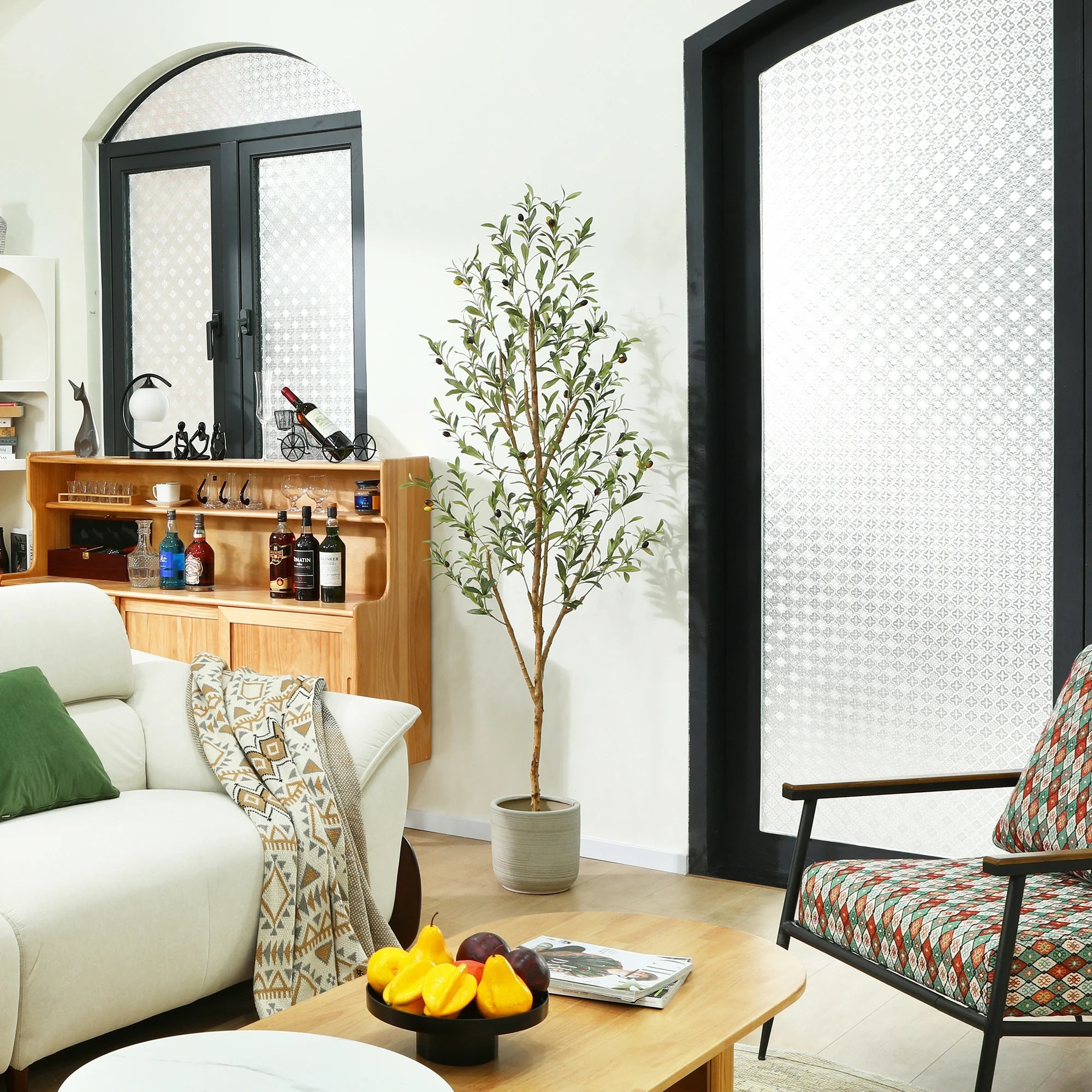 A cozy living room setup with a white sofa, patterned armchair, coffee table with fruit on it, a small tree in a pot, and shelving with bottles and glassware.