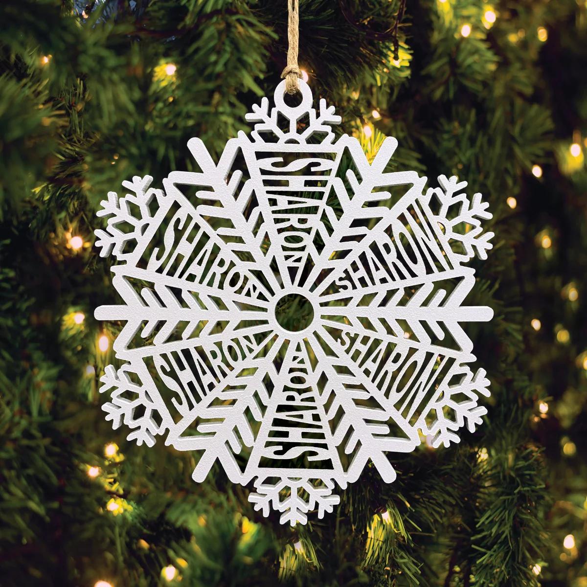 A white snowflake-shaped ornament with the name \“Sharon\“ integrated into the design hangs on a Christmas tree, surrounded by warm white lights.