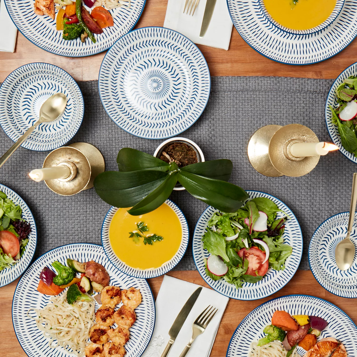 A dining setup features blue patterned plates with various dishes, including salad and soup, accompanied by gold cutlery and candlesticks. An orchid plant decorates the center.