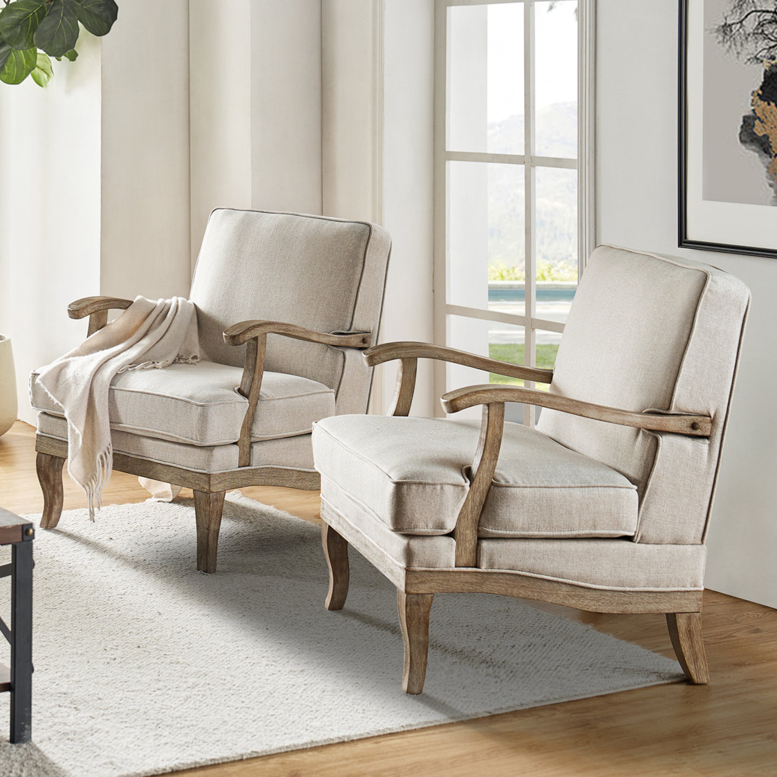 Two beige armchairs with wooden frames and cushioned seats, positioned in a light-filled room with a large window, an area rug, and a potted plant for a cozy, stylish setting.