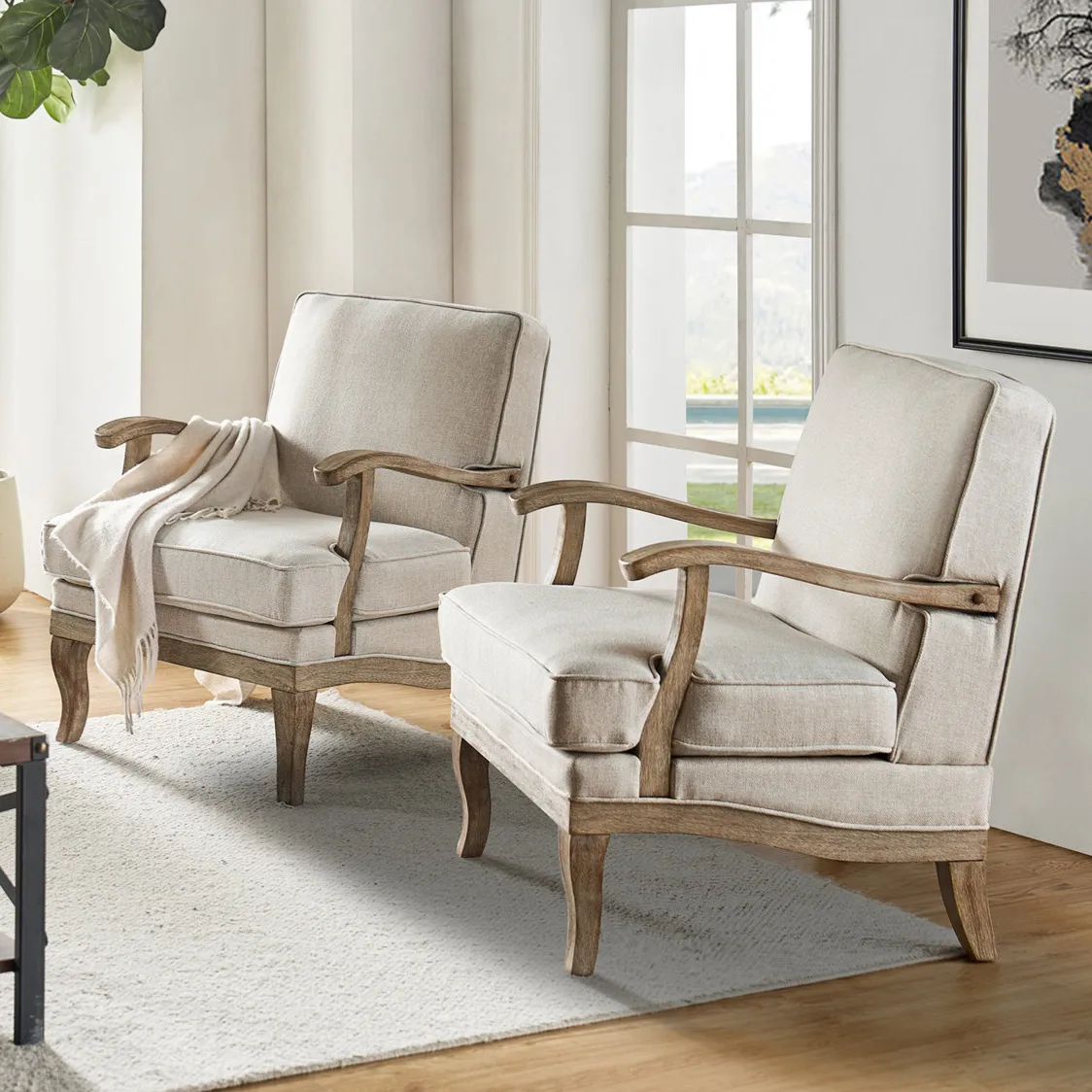 Two beige armchairs with wooden frames and cushioned seats, positioned in a light-filled room with a large window, an area rug, and a potted plant for a cozy, stylish setting.