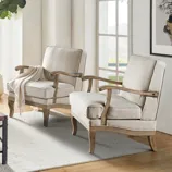 Two beige armchairs with wooden frames and cushioned seats, positioned in a light-filled room with a large window, an area rug, and a potted plant for a cozy, stylish setting.
