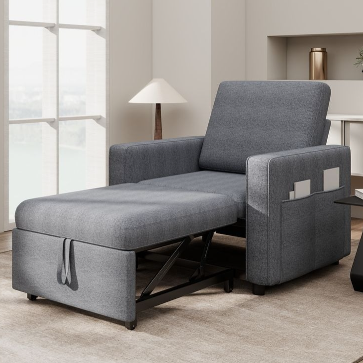 Convertible gray armchair with an extendable footrest, designed for lounging. Features a side pocket for storage and is placed in a modern room setting with a large window, lamp, and neutral furnishings.