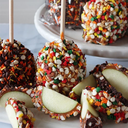 Caramel apples coated in various colorful sprinkles and chocolate chips, with some sliced to show the interior.