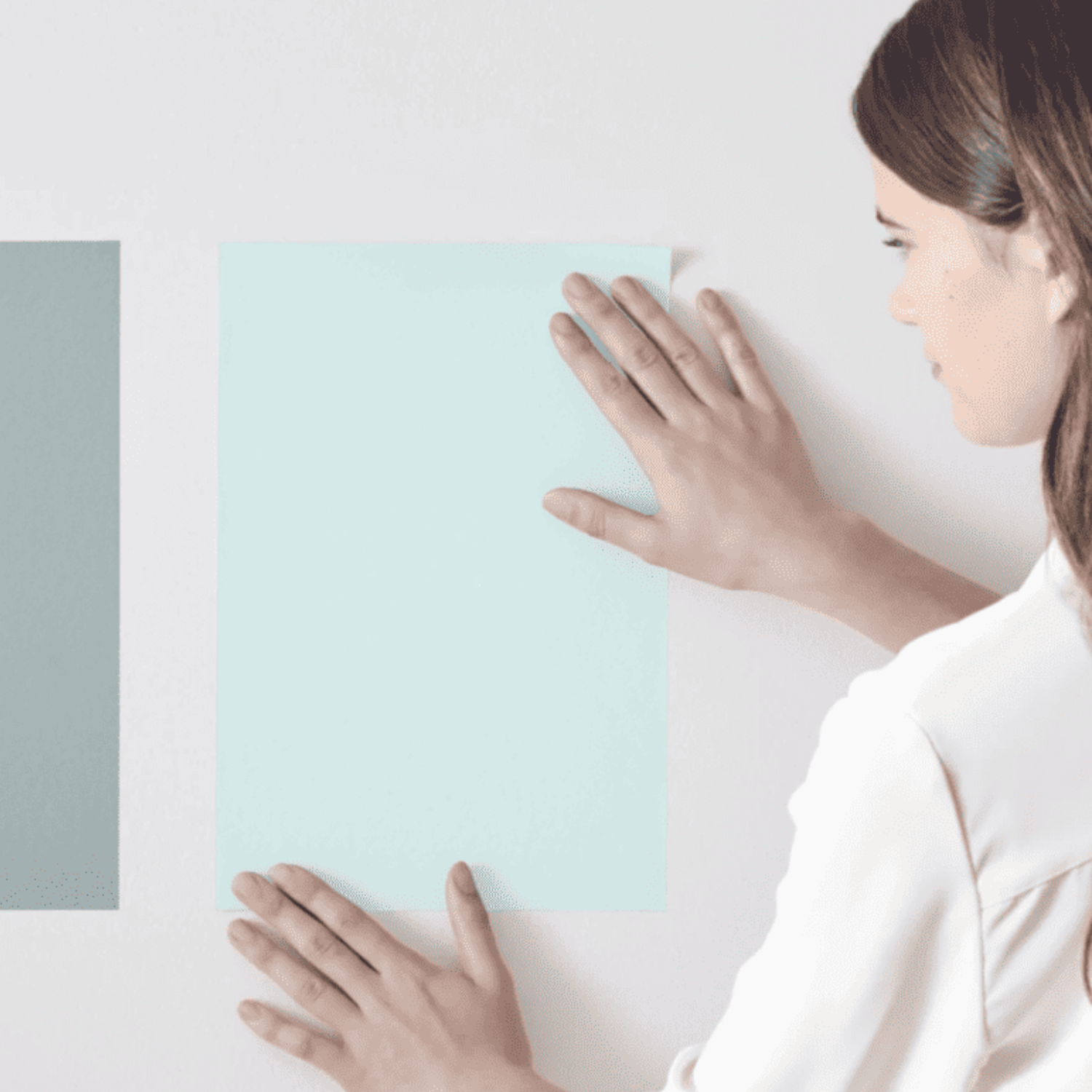 A person is aligning two pieces of colored paper, one light blue and the other teal, on a white wall.