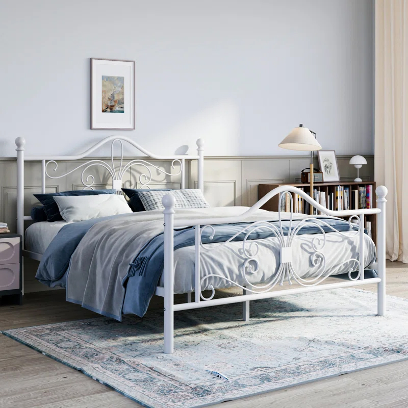 A white metal open-frame bed with ornate scrolling details on the headboard and footboard, set against a minimalist bedroom décor with blue and gray bedding, a soft rug, and a side table with books and a lamp.