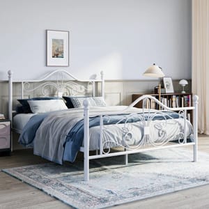 A white metal open-frame bed with ornate scrolling details on the headboard and footboard, set against a minimalist bedroom décor with blue and gray bedding, a soft rug, and a side table with books and a lamp.