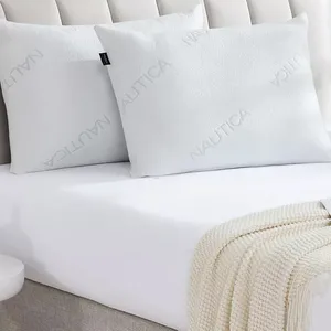 Two white pillows with the brand name \“Nautica\“ embossed on them are placed on a bed with a white sheet and a cream-colored knitted throw.