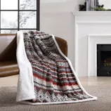 A cozy throw blanket with a red, brown, and white geometric pattern and a soft white backing is draped over a brown chair in a room with a fireplace and snowy window view.