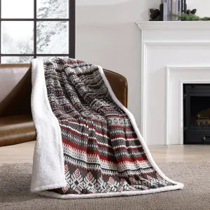 A cozy throw blanket with a red, brown, and white geometric pattern and a soft white backing is draped over a brown chair in a room with a fireplace and snowy window view.