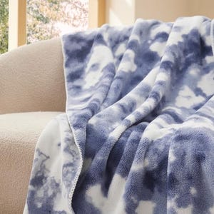 A plush chenille throw with a blue and white tie-dye pattern is draped over a cream-colored sofa near a window.