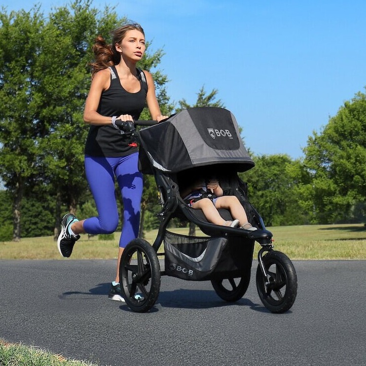 A woman is jogging with a child in a three-wheeled stroller.