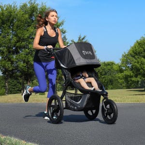 A woman is jogging with a child in a three-wheeled stroller.