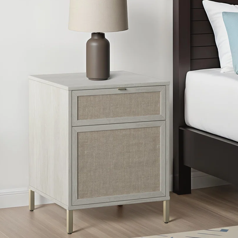 A nightstand with fabric-front drawers and a modern table lamp sits beside a bed. The nightstand has a light wood finish with slender metal legs.