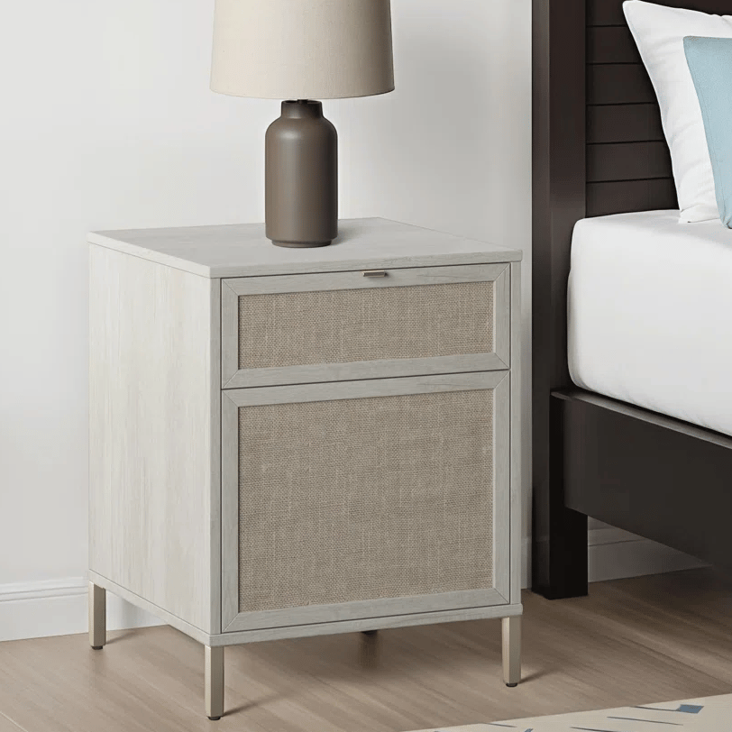 A nightstand with fabric-front drawers and a modern table lamp sits beside a bed. The nightstand has a light wood finish with slender metal legs.