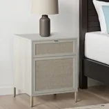 A nightstand with fabric-front drawers and a modern table lamp sits beside a bed. The nightstand has a light wood finish with slender metal legs.