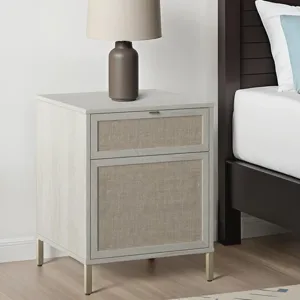 A nightstand with fabric-front drawers and a modern table lamp sits beside a bed. The nightstand has a light wood finish with slender metal legs.
