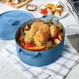 Blue Dutch oven filled with a roasted chicken and assorted vegetables, including carrots and peppers, on a white checkered cloth. A covered lid and side dish of roasted veggies are nearby on a marble countertop.