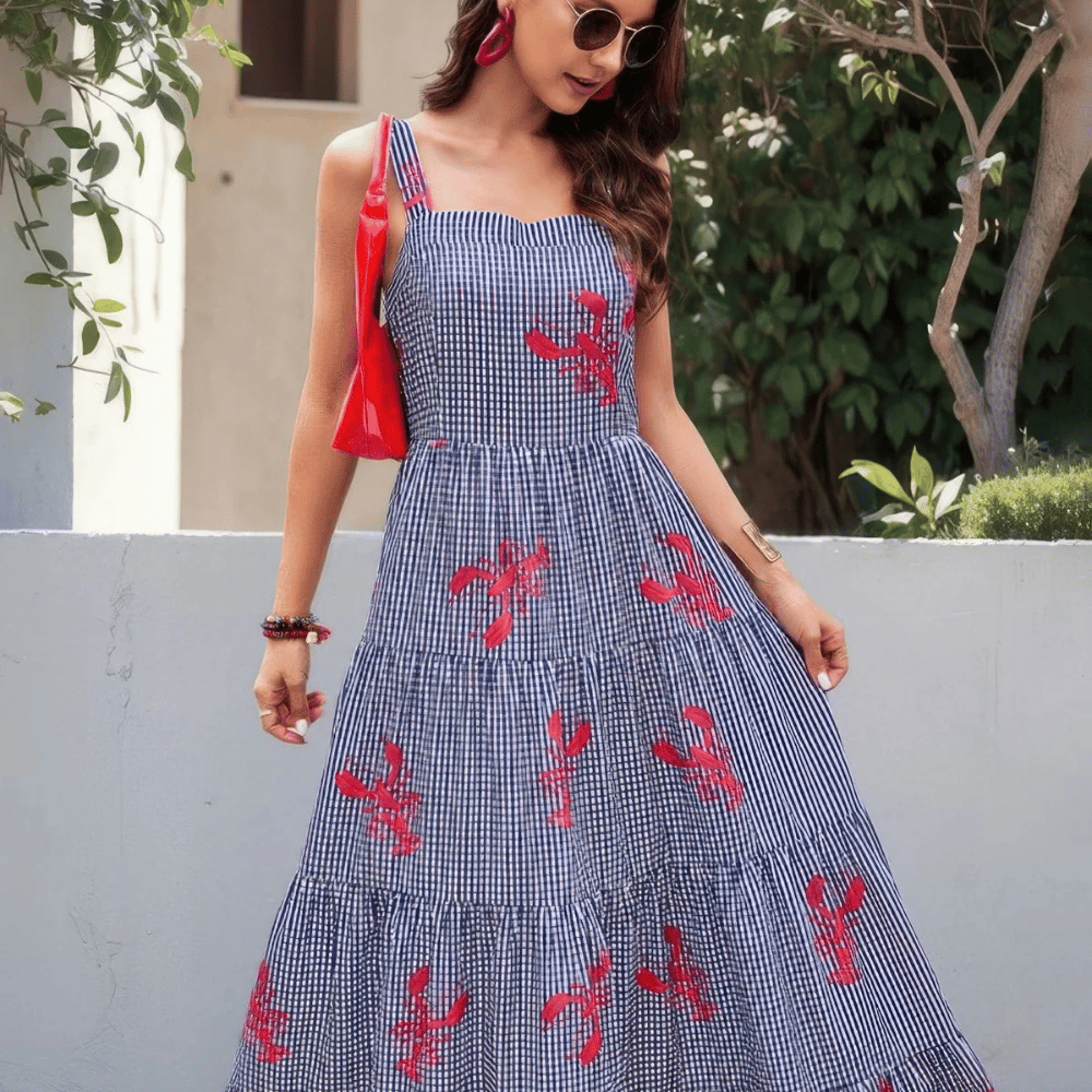 A woman wearing a blue and white checkered sundress with red floral patterns, accessorized with round sunglasses, red earrings, bracelets, and a red bag.