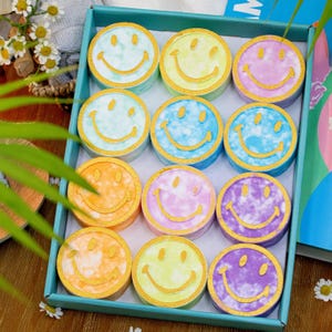 Twelve vibrantly colored shower steamers featuring smiley faces are arranged in a teal box. Each steamer has a unique tie-dye pattern, with shades including blue, yellow, purple, orange, and green, offering a cheerful and aromatic shower experience.