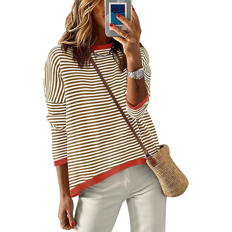 A woman wearing a striped long-sleeve top with red accents, white pants, a watch, and holding a phone. She carries a straw crossbody bag.